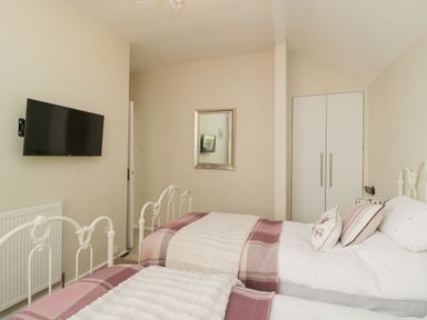 A bedroom with two white metal beds with pink and cream bedding a wall-mounted TV a mirror and a white wardrobe at Raven Cottage in Downderry