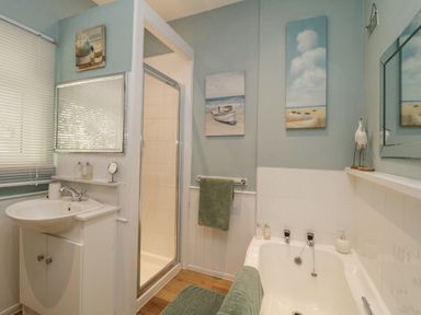 A bathroom with a sink, shower enclosure, bathtub, green towels, and paintings on the wall at Raven Cottage in Downderry