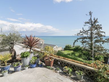 A patio with potted plants overlooking the sea and a grassy area at Raven Cottage in Downderry