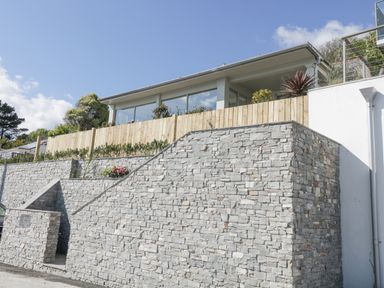 A modern house with large windows above a stone retaining wall and wooden fence at Raven Cottage in Downderry