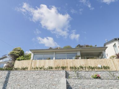 A modern house with large windows behind a wooden fence and stone retaining wall under a blue sky at Raven Cottage in Downderry