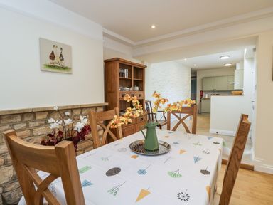 A dining area with a wooden table and chairs a vase with flowers on the table a wooden cabinet and kitchen area in the background at 2 Church Street in Chipping Norton
