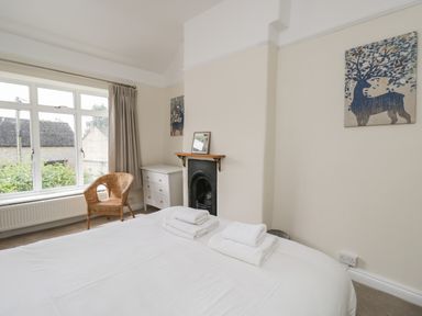 A bedroom with a bed with folded towels a wicker chair a white dresser a window and artwork at 2 Church Street in Chipping Norton