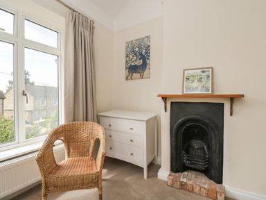 A wicker chair next to a window with beige curtains a white dresser a black fireplace and artwork on the walls at 2 Church Street in Chipping Norton