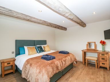 A bedroom with a bed featuring blue and gold pillows and a brown blanket a wooden nightstand and a wooden dressing table with a mirror and flowers at Palace Farmhouse in St Weonards