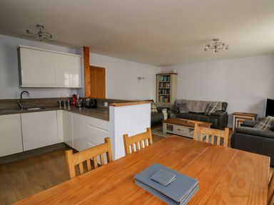 An open plan kitchen and living area with wooden dining table and chairs white kitchen cabinets grey sofas and a bookshelf at 1 St Marys Court in Tenby