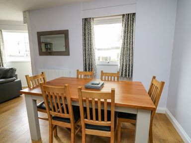 A dining area with a wooden table and six chairs near a window with patterned curtains at 1 St Marys Court in Tenby
