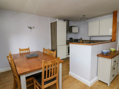 A dining area with a wooden table and six chairs next to a kitchen with white cabinets at 1 St Marys Court in Tenby