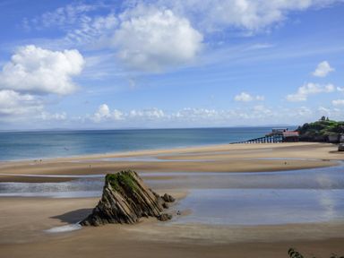 A sandy beach with a large rock formation near the shore and a pier with a building in the distance at 1 St Marys Court in Tenby