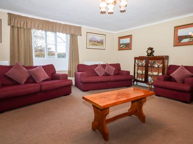 A living room with three red sofas a wooden coffee table a glass cabinet and framed pictures on beige walls at Twelve Oaks Farmhouse in Teigngrace near Newton Abbot