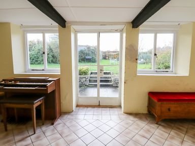 A room with a wooden organ and bench on the left a wooden chest with a red cushion on the right tiled floor and glass doors leading to a garden at Twelve Oaks Farmhouse Teigngrace near Newton Abbot