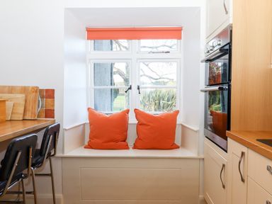 A kitchen window seat with two orange cushions and an orange blind next to a countertop with cutting boards and black chairs at Twelve Oaks Farmhouse Teigngrace near Newton Abbot