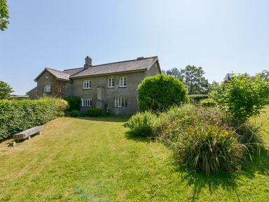 A grassy garden with bushes and a stone bench next to a two-story house at Twelve Oaks Farmhouse in Teigngrace near Newton Abbot