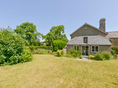 A grass garden with bushes and trees next to a small stone house with a slate roof at Twelve Oaks Farmhouse in Teigngrace near Newton Abbot