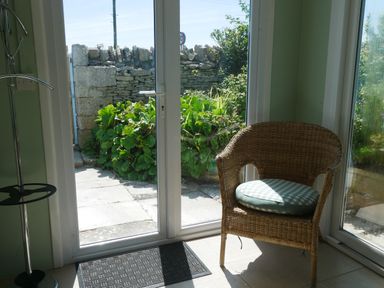 A wicker chair with a cushion next to glass doors opening to a garden with plants and stone wall at Fisherman's Cottage in Southwell
