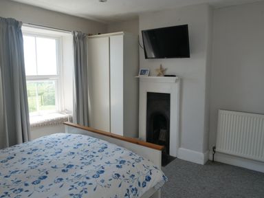 A bedroom with a bed covered in a blue and white floral duvet a window with gray curtains a white wardrobe a TV mounted above a small fireplace and a radiator on the wall at Fisherman's Cottage in Southwell