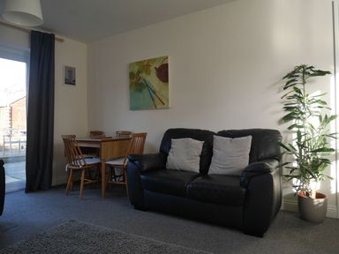 A living room with a black leather sofa and two cushions next to a dining table with wooden chairs and a potted plant at Cliffwalk Cottage in Southwell
