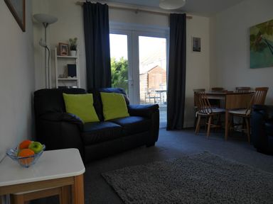 A living room with a black sofa with green cushions a wooden dining table with chairs and a small white side table with fruit at Cliffwalk Cottage in Southwell