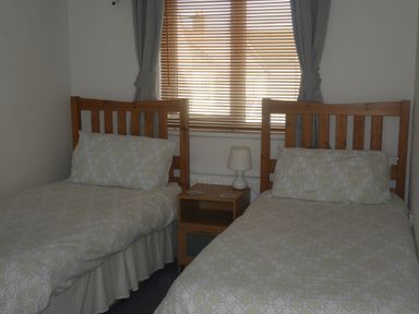 A bedroom with two single beds with wooden headboards a nightstand with a lamp and a window with blinds and curtains at Cliffwalk Cottage in Southwell