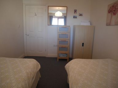 A bedroom with two beds a white door a wooden chest of drawers a mirror a wardrobe and a painting at Cliffwalk Cottage in Southwell