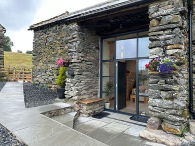 Stone exterior of a building with large glass doors and hanging flower baskets at Meadow Syke Barn in Bampton Cumbria