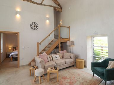 A living room with a beige sofa, green armchair, small wooden tables, stairs with wooden railing and glass panels at Meadow Syke Barn in Bampton Cumbria