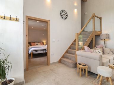 A living area with stairs a beige sofa and a bedroom visible through a doorway at Meadow Syke Barn in Bampton Cumbria