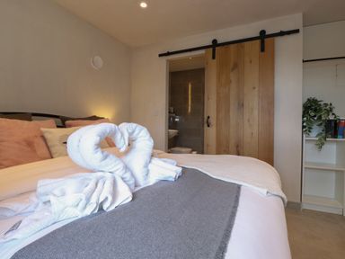 A bedroom with a bed with towels folded into swan shapes a sliding wooden door and a small shelf with books and a plant at Meadow Syke Barn in Bampton Cumbria
