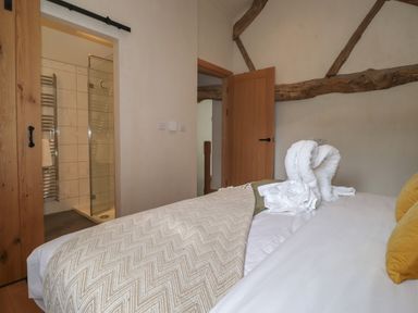 A bedroom with a bed covered by a beige blanket and two yellow pillows next to a bathroom with a glass shower door at Meadow Syke Barn in Bampton Cumbria
