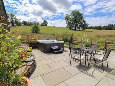 An outdoor patio with a hot tub and a dining table with four chairs overlooking a grassy field at Meadow Syke Barn in Bampton Cumbria