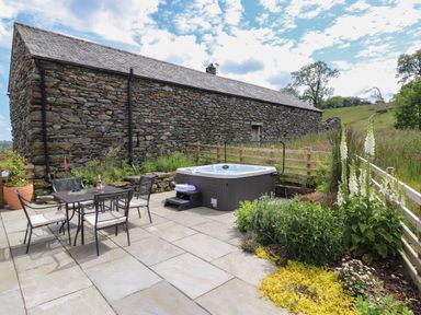 An outdoor patio with a dining table and chairs and a hot tub near a stone building with a garden at Meadow Syke Barn in Bampton Cumbria