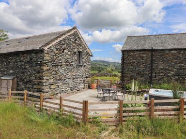 An outdoor patio area between two stone buildings with a table and chairs a hot tub and a wooden fence surrounding a garden at Meadow Syke Barn in Bampton Cumbria