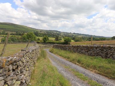 A gravel pathway between stone walls and fields with trees and hills in the background at Meadow Syke Barn in Bampton Cumbria
