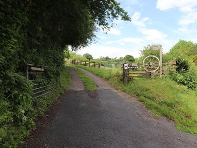 A paved driveway leading to a gravel path with a wooden fence and a sign reading Meadowsyke in a rural setting at Meadow Syke Barn in Bampton Cumbria