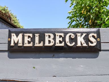A black wooden sign with white letters spelling Melbecks mounted on a grey wooden fence with greenery and blue sky behind at Melbecks in Abergele