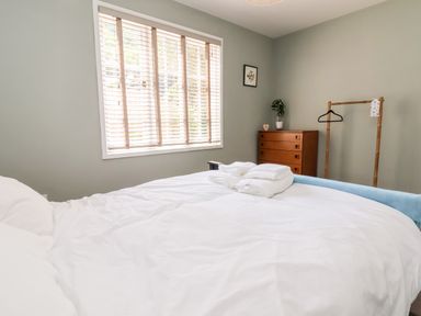 A bedroom with a bed covered in white sheets folded towels on top a window with blinds a wooden chest of drawers a plant a clothes hanger and a framed picture on the wall at Melbecks in Abergele
