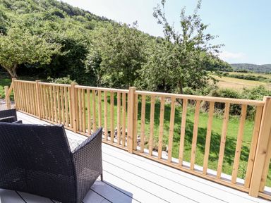 A wooden deck with dark woven chairs and wooden railing overlooking grass trees and hills at Melbecks in Abergele