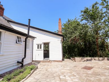 An outdoor stone patio area beside a white wooden house with a black drainpipe and a garden bed with plants near trees at Melbecks in Abergele