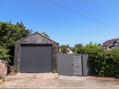 A garage with a closed black roller door next to a wooden gate with Melbecks sign and greenery around in Abergele