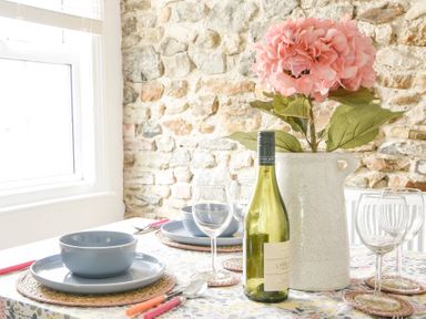 A dining table set with plates bowls wine glasses and cutlery with a bottle of wine and a vase with pink flowers against a stone wall at River Lym Cottage Lyme Regis
