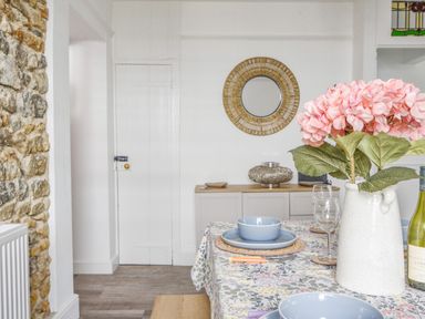 A dining area with a floral tablecloth a white vase with pink flowers blue bowls and plates wine glasses and a stone wall at River Lym Cottage Lyme Regis