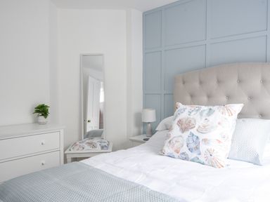 A bedroom with a bed with seashell pillows a nightstand with a lamp a chest of drawers with a plant and a full length mirror at River Lym Cottage in Lyme Regis