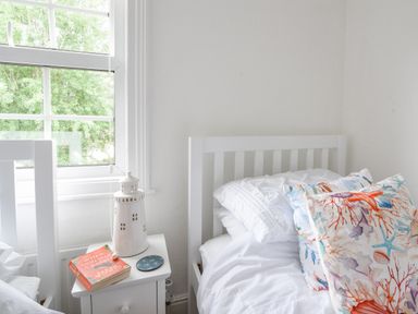 A bedroom corner with a white bed and colorful pillows and a white nightstand with a book and lighthouse decoration at River Lym Cottage Lyme Regis