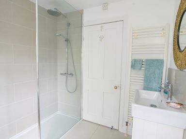 A bathroom with a shower area a white door a sink with a mirror and towels on a radiator at River Lym Cottage in Lyme Regis