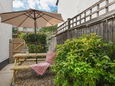An outdoor seating area with a wooden picnic table umbrella and a pink blanket with a bottle and two glasses on the table at River Lym Cottage in Lyme Regis