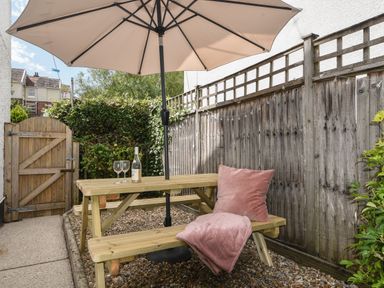 An outdoor patio area with a wooden picnic table and bench under a large beige umbrella with two wine glasses and a wine bottle on the table and a pink cushion and blanket on the bench at River Lym Cottage in Lyme Regis