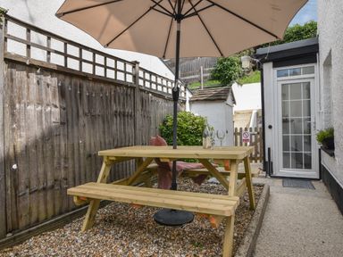 An outdoor seating area with a wooden picnic table umbrella and a bottle with two glasses near a white door at River Lym Cottage in Lyme Regis