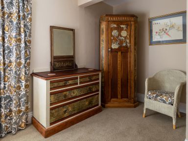 A corner of a room with a wooden dresser with a mirror a wooden corner cabinet a wicker chair with patterned cushion and floral curtains at The Lodge in Hartlington near Grassington