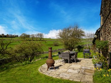 An outdoor patio with a dining table and chairs a chiminea and a barbecue grill next to a stone building with trees and green fields in the background at The Lodge in Hartlington near Grassington