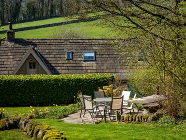 A garden patio with a round table and chairs next to a house with a tiled roof at The Lodge Hartlington near Grassington